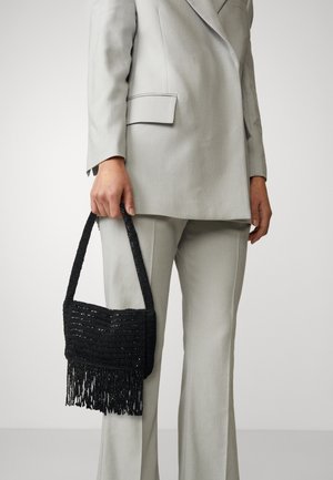 Person wearing light gray tailored suit holding black woven handbag with fringe detail by their side.