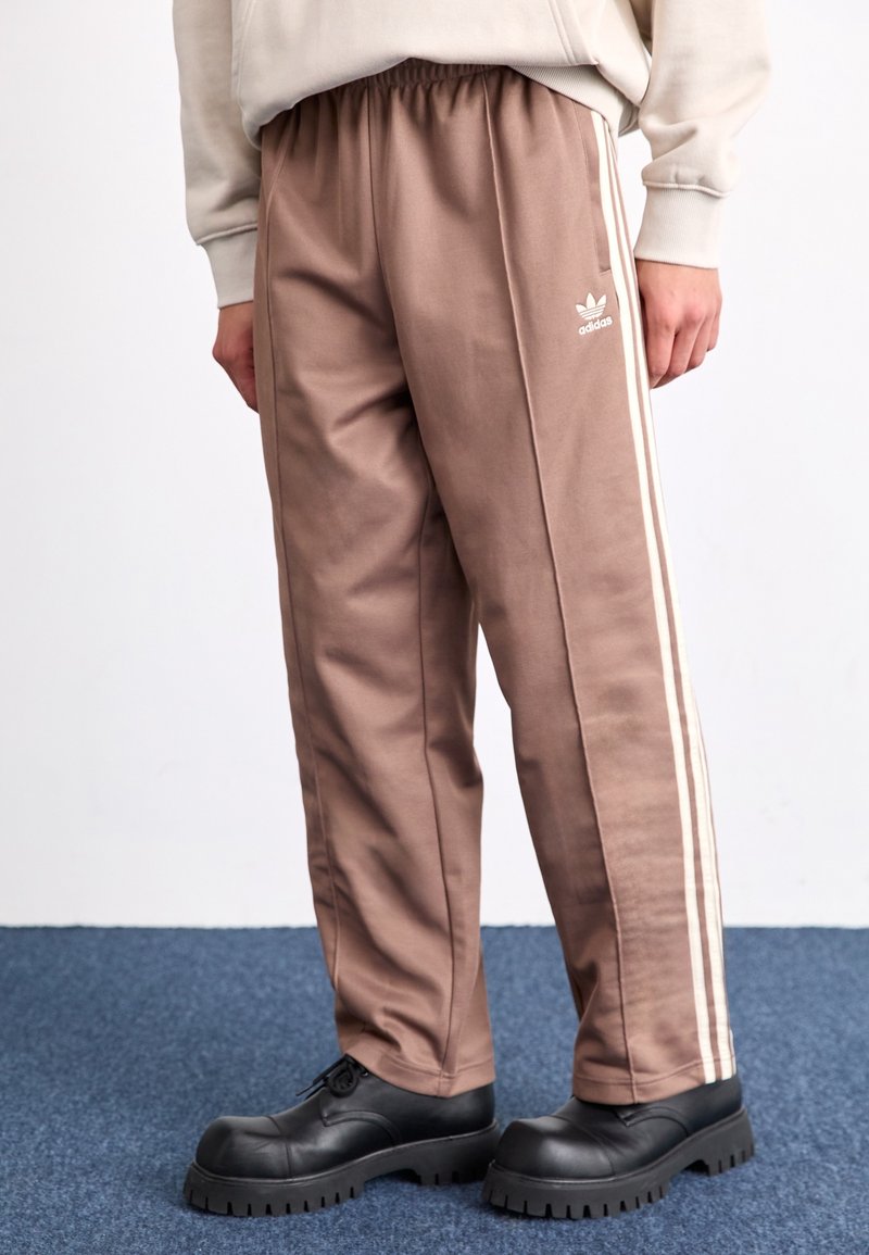 Bruna träningsbyxor med elastisk midja, vita randiga detaljer längs sidorna och en liten vit logotyp. Slät tyg med rak passform.