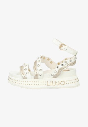 Białe sandały na platformie z ozdobnymi ćwiekami i dżetami. Charakteryzują się grubą podeszwą oraz paskiem na kostkę z klamrą. Nazwa marki jest widoczna po boku.