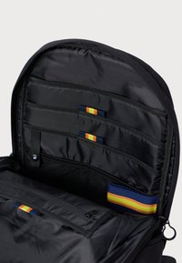 Interior de mochila negra con tres compartimentos, bolsillos de malla y correas de colores con acentos en rojo, amarillo y azul. Cierre de cremallera en la parte superior.
