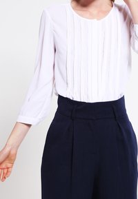 White blouse with vertical pleats, round neckline, and three-quarter sleeves, paired with high-waisted navy trousers featuring front pleats.