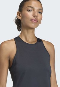 Eine Frau trägt ein schwarzes ärmelloses Sportoberteil mit einem kleinen Logo. Sie blickt nach vorne, ihr Haar ist nach hinten gebunden, vor einem schlichten hellen Hintergrund.