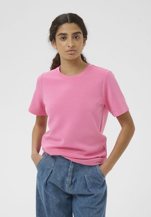 Mladá žena s kučeravými vlasmi, oblečená v ružovej tričku s krátkym rukávom a modrých džínsach s vysokým pásom, stojaca s rukami v vreckách.