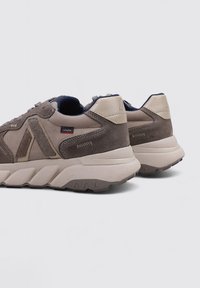 Une paire de baskets en daim beige et marron avec de épaisses semelles en caoutchouc et un intérieur bleu marine, vue de l'arrière et de côté sur un fond uni.