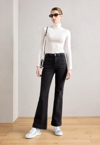Black flared jeans with front pockets, worn with a white turtleneck top. Accessories include black sunglasses and white sneakers.