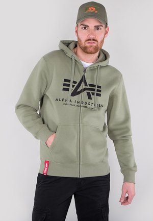 Zelena zip-up obična s kapuljačom, crnim logotipom, džepovima u stilu kengura i crvenom oznakom na rubu. Tkanina izgleda meko i teksturirano.