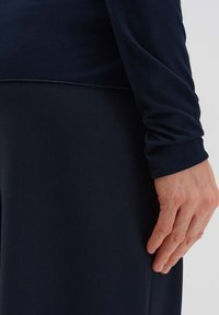 Langarm, navyblaues Oberteil aus weichem Material, das ein tailliertes Design mit einem runden Halsausschnitt und Umschlagärmeln zeigt. Die Hand ruht an der Seite.