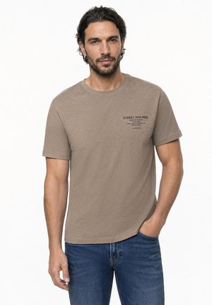 Homme aux cheveux foncés et à la barbe, portant un t-shirt marron avec un petit texte sur la poitrine et un jean bleu, debout avec une main dans la poche.