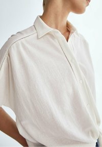 Weißes Leinenhemd mit kurzen Ärmeln, Kragen, Knopfvorn, und dekorativer Stickerei entlang der Schulter nähte.