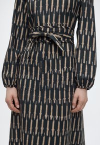 Μαύρο φόρεμα με κατακόρυφο μπεζ μοτίβο από πινελιές, μακριά φουσκωτά μανίκια, μπροστινές τσέπες και δεμένη υφασμάτινη ζώνη στη μέση.