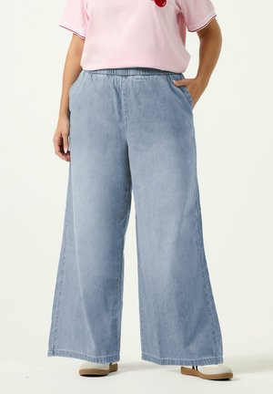 Person trägt hellblaue Jeans mit weitem Bein, rosa kurzärmeliges Hemd, weiße Schuhe mit braunen Sohlen, eine Hand in der Hosentasche, steht vor weißem Hintergrund.