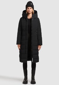Long black puffer coat with a hood, featuring horizontal quilted sections, two side zippers, and a logo tag at the hem. Worn with a black beanie.