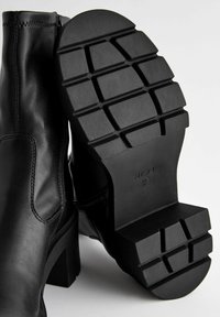 Black leather ankle boots with a high block heel and a ribbed rubber sole featuring a grid pattern. Side zipper closure for easy wear.
