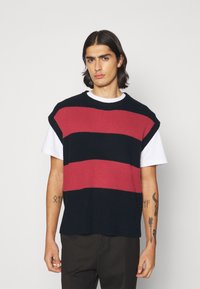Gilet en tricot avec de larges rayures rouges et noires, manches courtes, texture côtelée et col haut, superposé sur un t-shirt blanc.