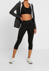 Brassière de sport noire, sweat à capuche gris zippé et leggings capri noirs avec des bandes latérales blanches. Complété par des chaussures de sport gris clair.