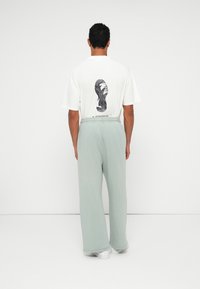 Herren T-Shirt aus weißer Baumwolle mit einem schwarzen Grafikdruck auf der Rückseite. Kombiniert mit hellgrünen, locker sitzenden Jogginghosen und weißen Sneakern.