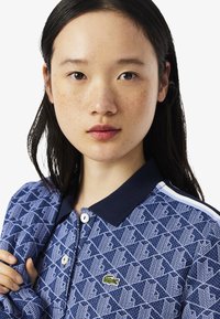 Polo bleu à motifs avec un col marine, des accents blancs et un petit logo vert. Le tissu présente un design texturé avec des formes géométriques.