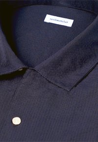 Chemise à col texturée foncée avec des boutons blancs et une étiquette blanche portant l'inscription "seidensticker" à l'intérieur du col.