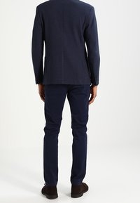 Granatowa teksturowana marynarka z pojedynczym rozcięciem z tyłu, noszona na ciemnych spodniach, widok od tyłu, w zestawieniu z brązowymi butami wsuwanymi. Gładka tkanina i dopasowany krój.