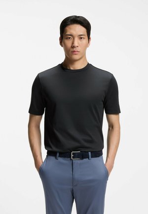 Uomo in piedi, rivolto in avanti con le mani nelle tasche di pantaloni azzurri, indossa una maglietta nera a maniche corte e una cintura scura su uno sfondo bianco.