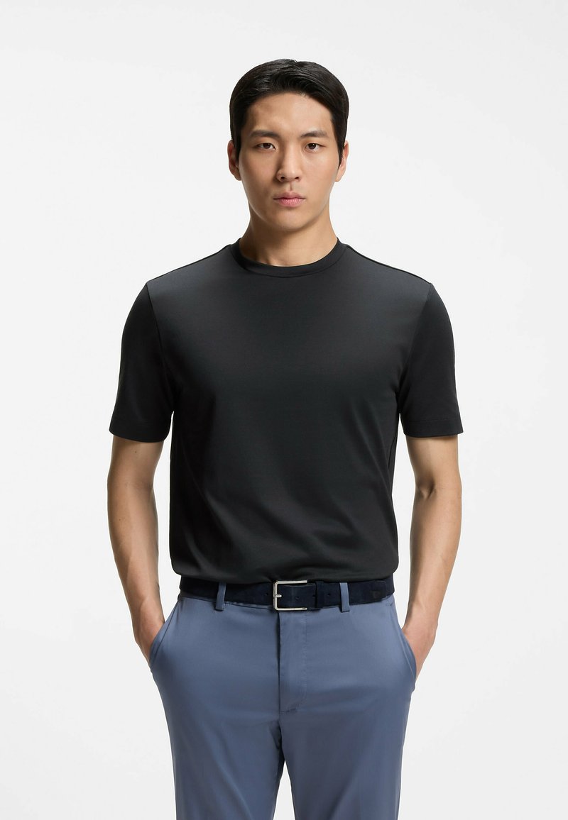 Un homme se tient face à l'avant, les mains dans les poches de son pantalon bleu clair, portant une chemise noire à manches courtes et une ceinture foncée sur un fond blanc.