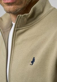 Beige zip-up jakke lavet af blødt stof med en opretstående krave. Har et lille navy broderet logo på venstre bryst.