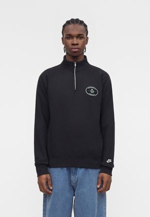Ung mand med fletninger iført en sort Nike quarter-zip sweatshirt med logopatches, kombineret med løstsiddende blå jeans, mod en ensfarvet baggrund.