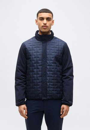 Jeune homme portant une veste matelassée bleu marine avec des manches unies, debout devant un fond clair uni, faisant face à l'avant.