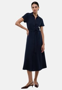 Robe chemise bleu marine avec des boutons et des manches courtes, ceinturée à la taille. Texture lisse, longueur genoux, associée à des chaussures à talons marron.