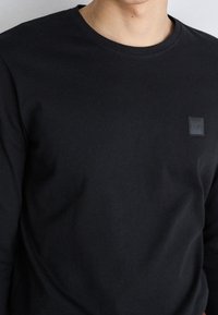 Black long-sleeve t-shirt made of cotton with a round neckline and a small square BOSS logo patch on the left chest. Smooth texture.