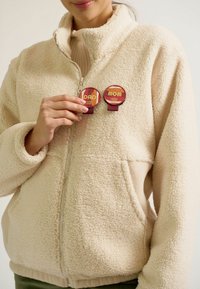 Beige Fleece-Jacke mit Reißverschluss und strukturiertem Oberflächen, versehen mit runden Anstecknadeln, die in warmen Farben "DAD" und "MOM" beschriftet sind.