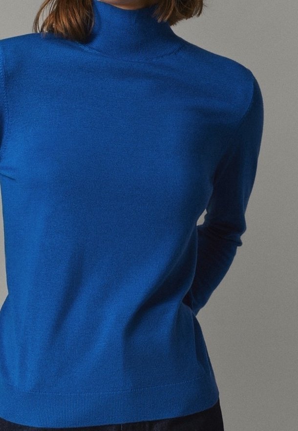 Femme portant un pull à col roulé bleu uni à manches longues, les mains dans le dos sur un fond neutre.