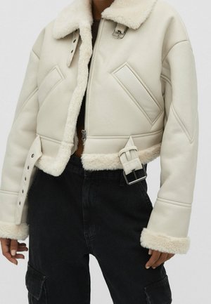 Kremowa krótka kurtka z kożuszkiem, ozdobiona klamrą i zamkami, noszona z czarnymi, wysokim stanem spodniami typu cargo.