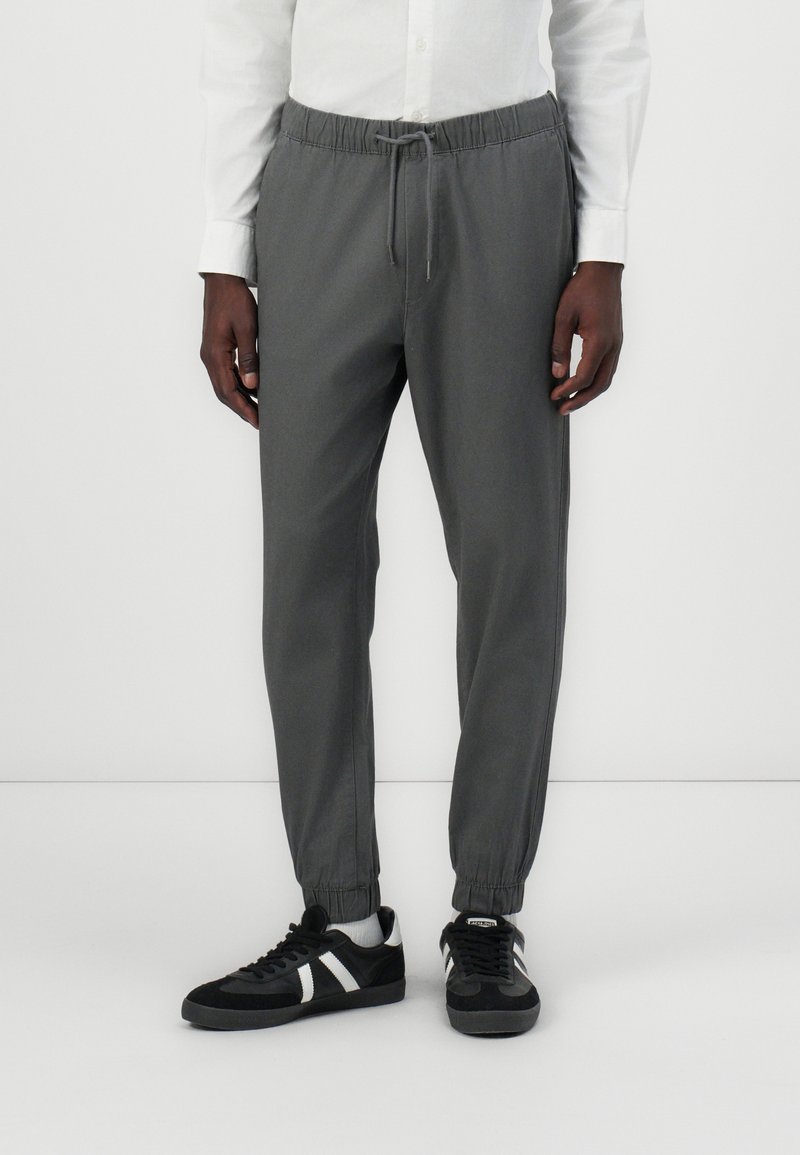 Homme portant un pantalon de jogging gris foncé avec des chevilles élastiques et une taille à cordon, des baskets noires à rayures blanches et une chemise blanche.