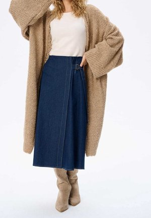 Femme portant un cardigan en maille beige oversize, un haut blanc, une jupe portefeuille bleu foncé avec boucle, et des bottes hautes en daim beige, debout devant un fond blanc.