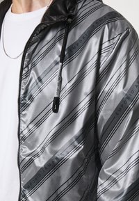 Silver zip-up jacka med en slät, glänsande textur och diagonala svarta ränder samt "Emporio Armani"-logotyp på framsidan. Svart huva.