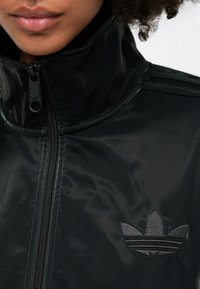 Juodas sportinis švarkas su aukštu apykakle, blizgiu paviršiumi, pilnu priekiu užtrauktuku ir išsiuvinėtu logotipu, kuriame yra trys lapų formos akcentai.