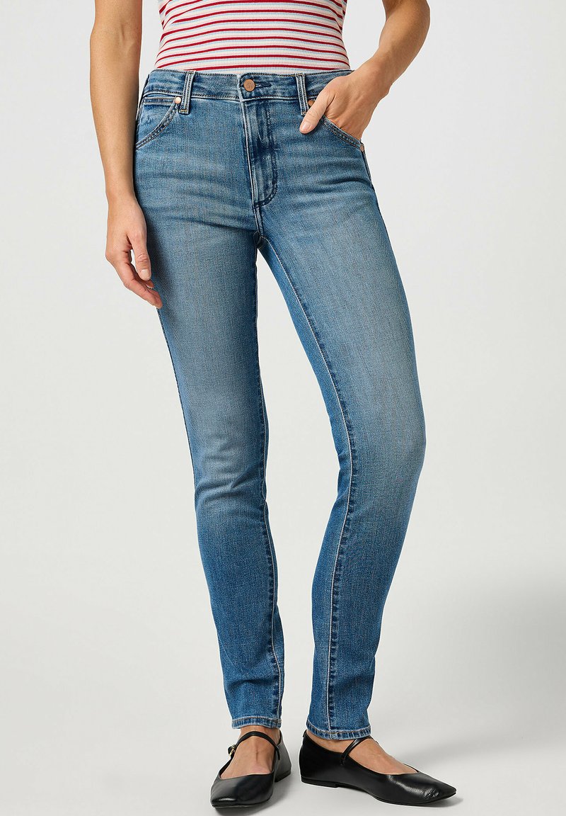 Jeans ajustados de mezclilla azul, con una cintura de altura media, cinco bolsillos, un ligero desgaste y un corte clásico. Combinados con zapatos planos negros.