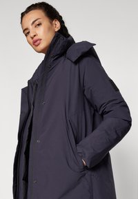 Giacca puffer blu scuro con colletto alto, chiusura a zip e bottoni, tasche laterali e una texture liscia e opaca.
