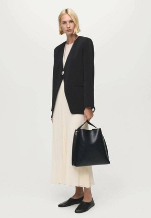 Blazer negro sobre un vestido crema, llevando un bolso tote de cuero negro. Zapatos planos negros. Diseño simple con líneas limpias y mínimos acentos.