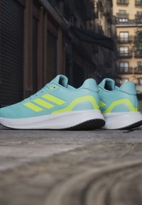 Baskets de sport bleu clair avec des accents jaune, matériau supérieur lisse, épaisse semelle intermédiaire blanche et semelle extérieure noire texturée. Logo à trois bandes visible.