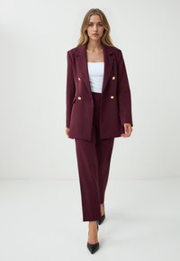 Bordeaux dubbelgeknopte blazer en bijpassende broek, met gouden knopen en een gladde textuur, gecombineerd met een witte top en zwarte hoge hakken.
