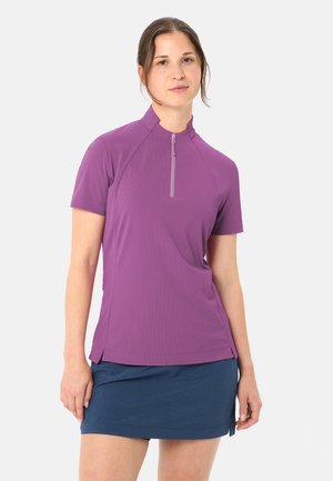 Giovane donna con indosso una maglietta viola a maniche corte con cerniera e una gonna blu scuro, in piedi con un'espressione neutra su uno sfondo bianco.