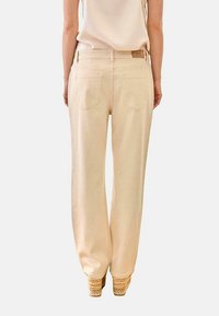 Mujer con pantalones anchos de talle alto color beige con bolsillos traseros y top sin mangas beige, de pie sobre sandalias plataformas tejidas.