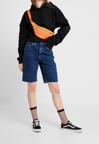 Černá mikina s kapucí, denimové šortky, oranžová crossbody taška, síťované ponožky a černé tenisky, což vytváří neformální, vrstvený vzhled.
