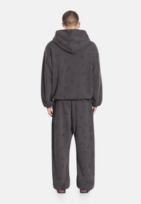 Mężczyzna ubrany w ciemnoszarą bluzę z kapturem oraz pasujące, luźne spodnie z subtelnymi nadrukami owadów, stojący tyłem na białym tle.