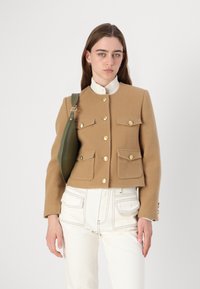 Brun uldjakke med guldknapper, fire forreste lommer og en høj hvid krave, parret med cremefarvede jeans og en grøn taske.