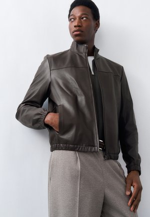 Jeune homme portant une veste en cuir marron foncé à poignets élastiques et un pantalon gris, debout avec une main dans la poche contre un mur blanc.