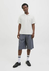 Kortærmet, hvid polo-shirt med struktureret stof, parret med grå knæ-lange shorts. Sorte snørebåndssko og hvide sokker med sorte striber.