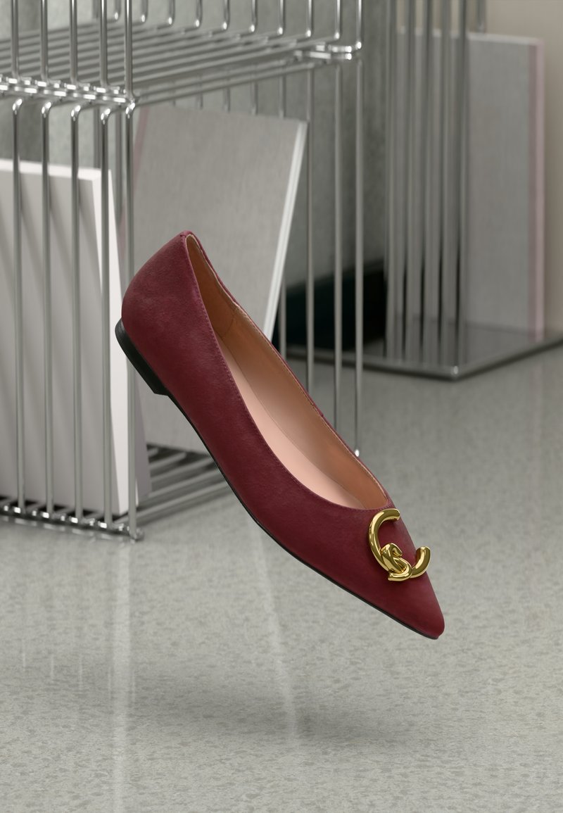 Sapato plano de couro bordô com bico fino, apresentando um detalhe em ouro na frente e um salto baixo preto. Textura suave, design minimalista.